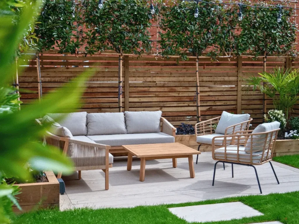 Cozy outdoor living space featuring natural wooden furniture, patio decking, and a slatted horizontal privacy fence with trained pleached trees.